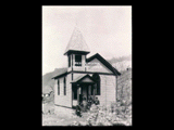 Nuttalburg church. Frame church with Gothic windows with seven people standing or sitting at the entrance. A stone wall is in front at right.